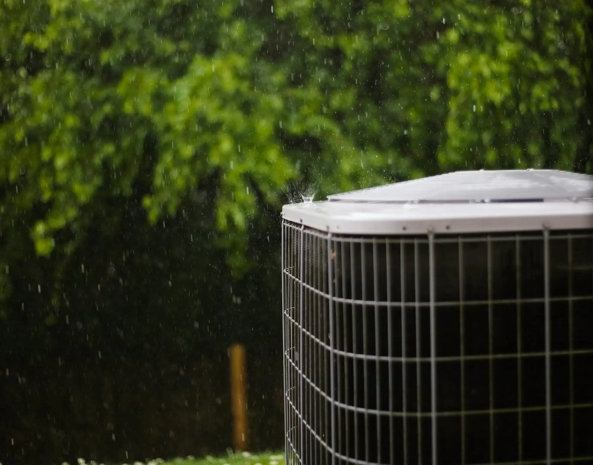 Outdoor air conditioning condenser in a summer rain shower, green trees behind it