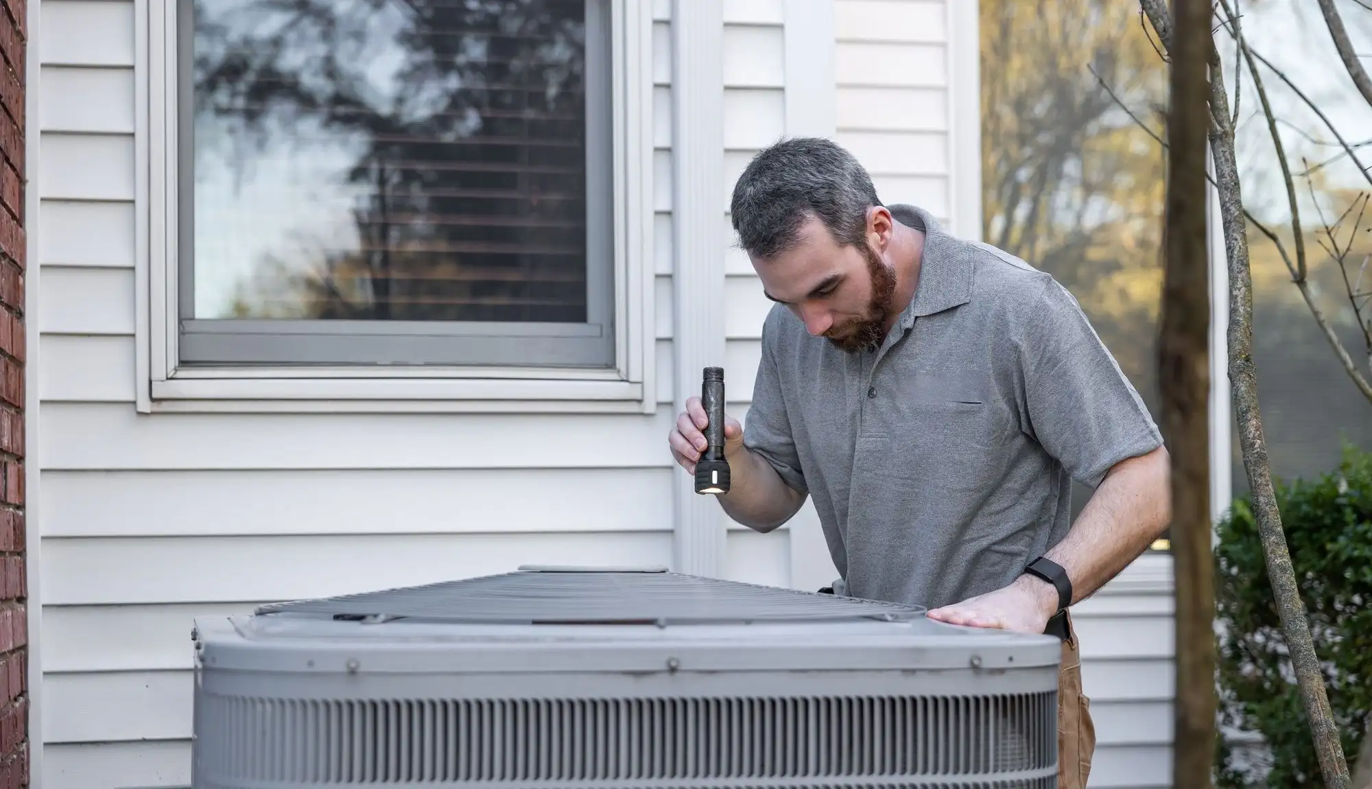 Technician shining a flashlight into the top of an outdoor heat pump unit beside a house