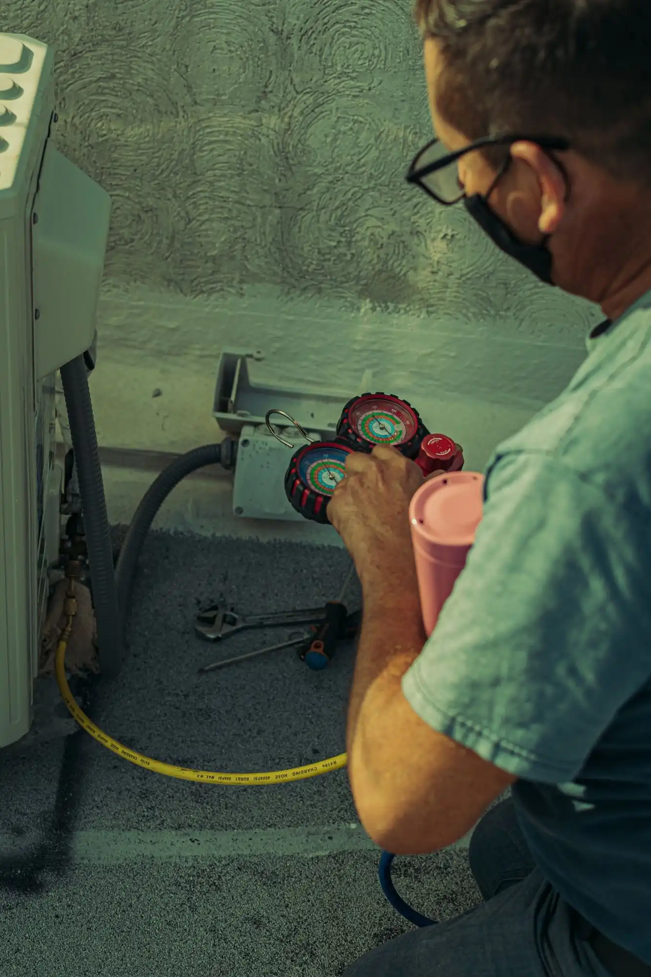 Technician crouched beside an outdoor AC unit against a stucco wall, reading a refrigerant gauge set connected to the service ports
