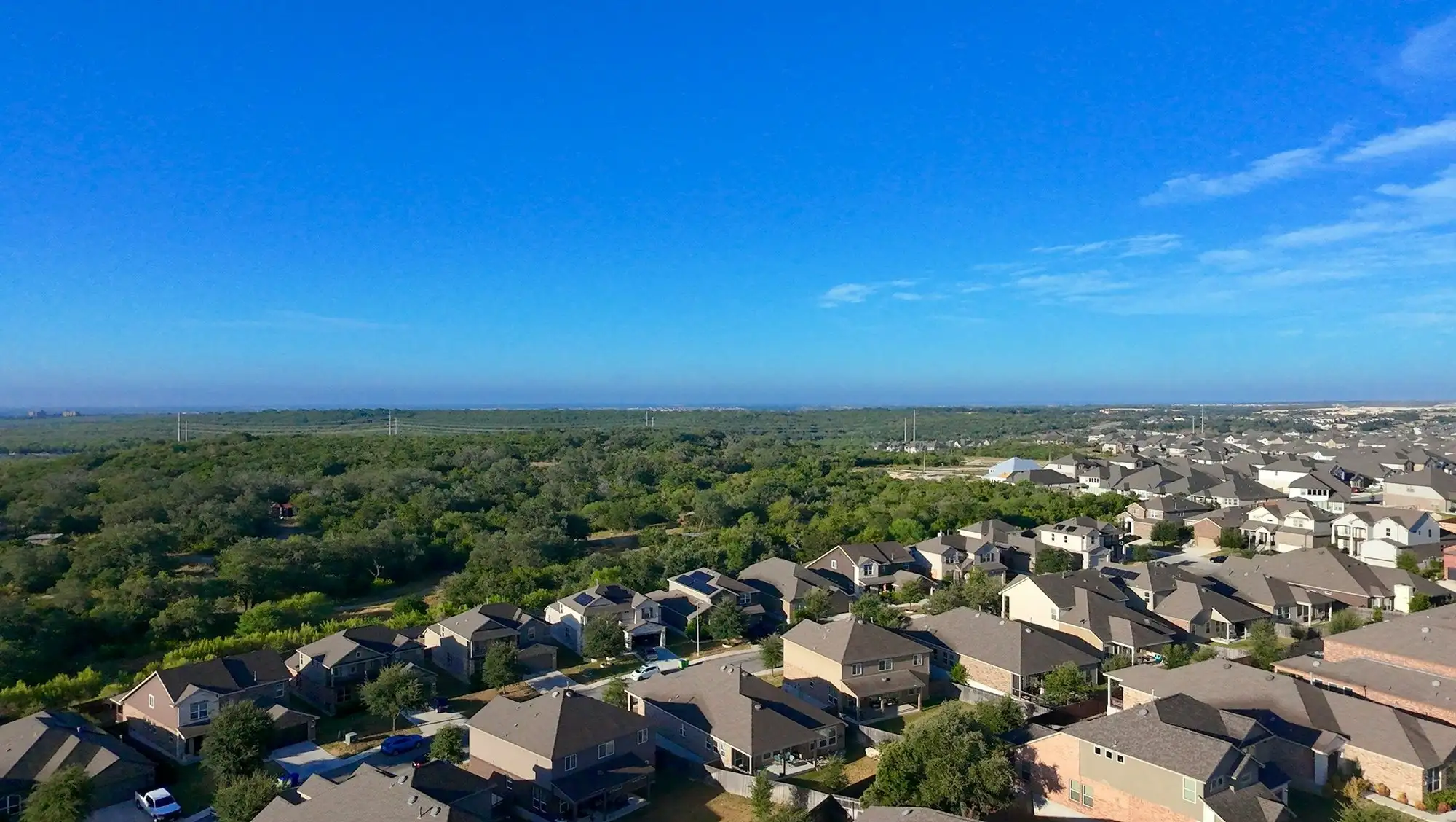 Rooftops of a new subdivision at the edge of dense oak and cedar woodland under a clear Texas sky
