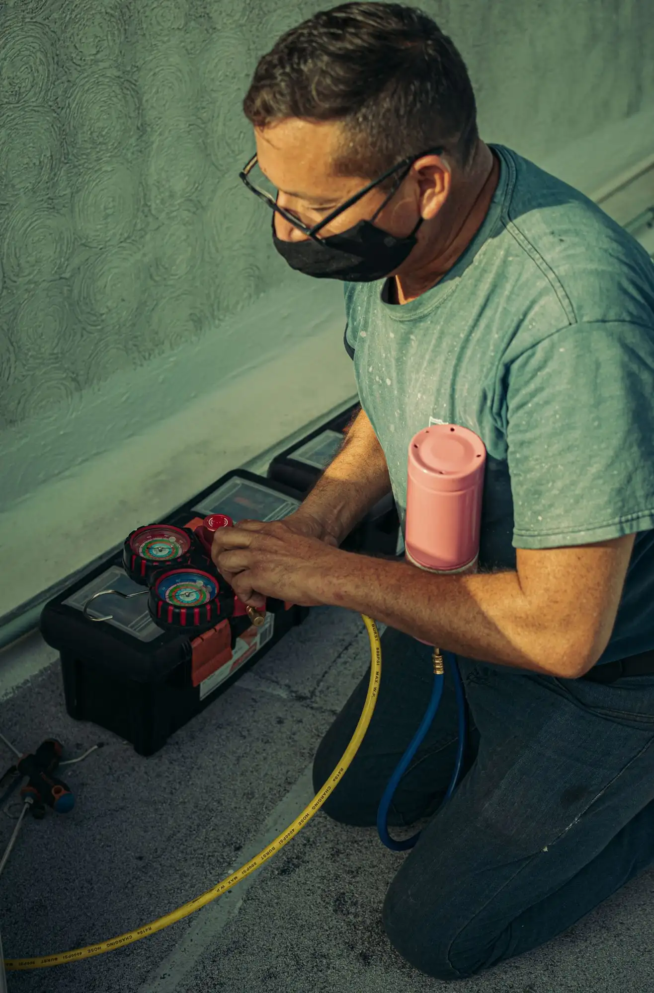 Technician kneeling on a concrete pad, setting up refrigerant gauges on an open toolbox
